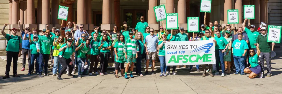 Workers in green AFSCME shirts outside of City Hall holding up a banner that says 'Say Yes to AFSCME 189'